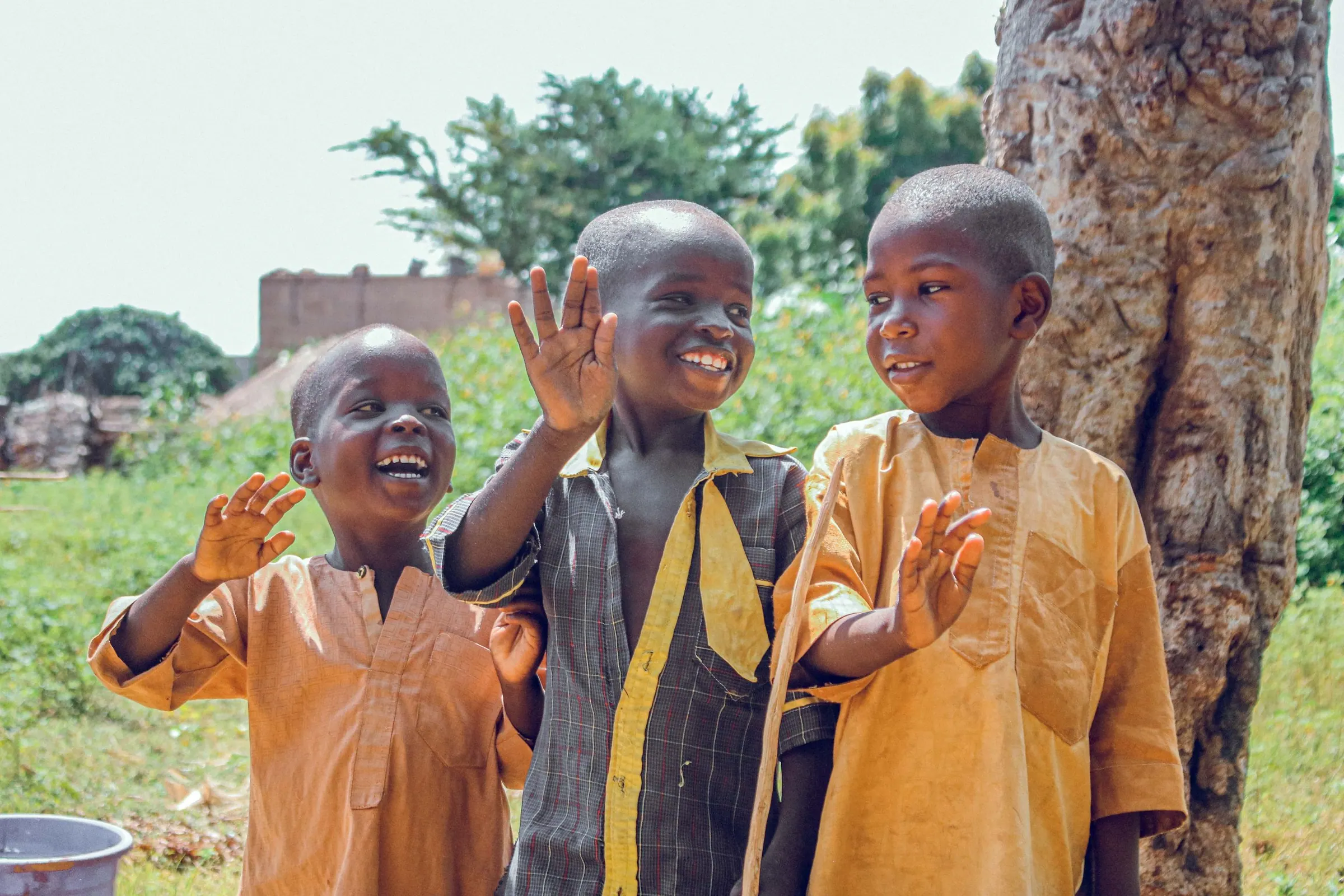 Three boys smiling and waving outdoors in a Nigerian village